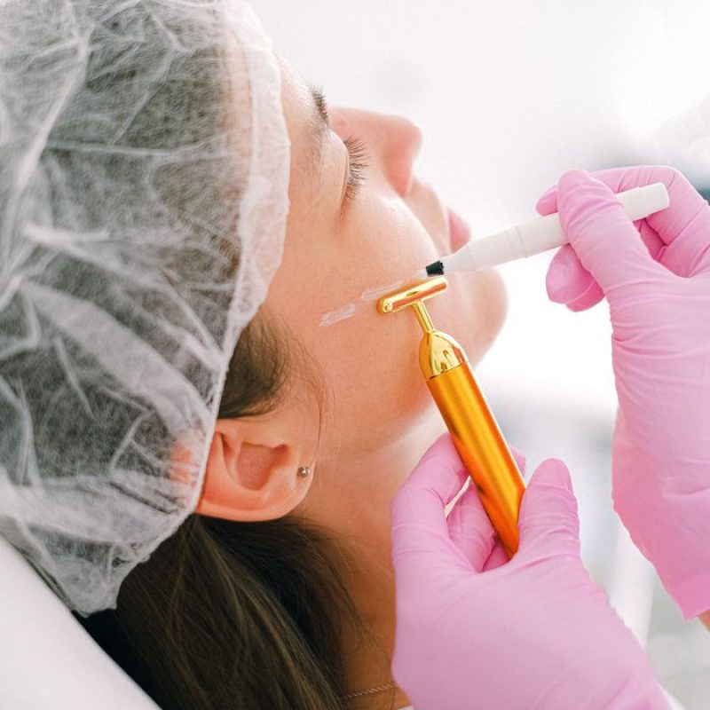 A cosmetologist performs a facial treatment using a golden massage roller and marker.