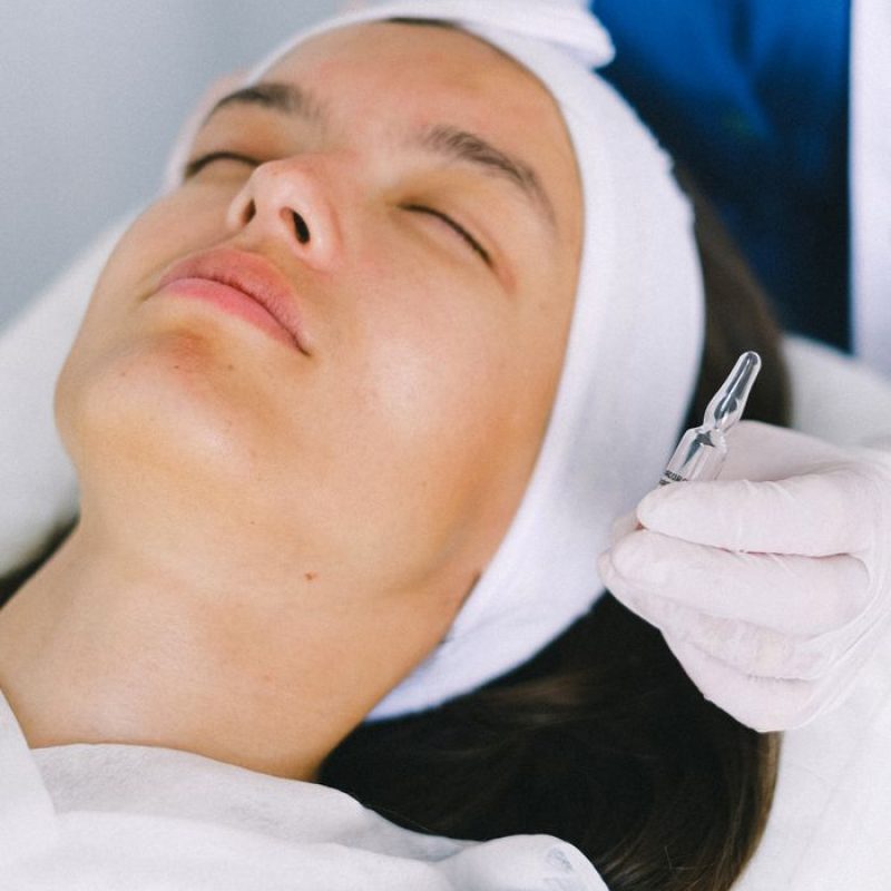Young relaxed brunette lying with eyes closed on lounge chair near cosmetologist in modern beauty clinic